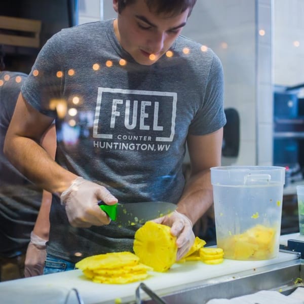 A Fuel Counter employee in a gray shirt and gloves slices fresh pineapple on a white cutting board.