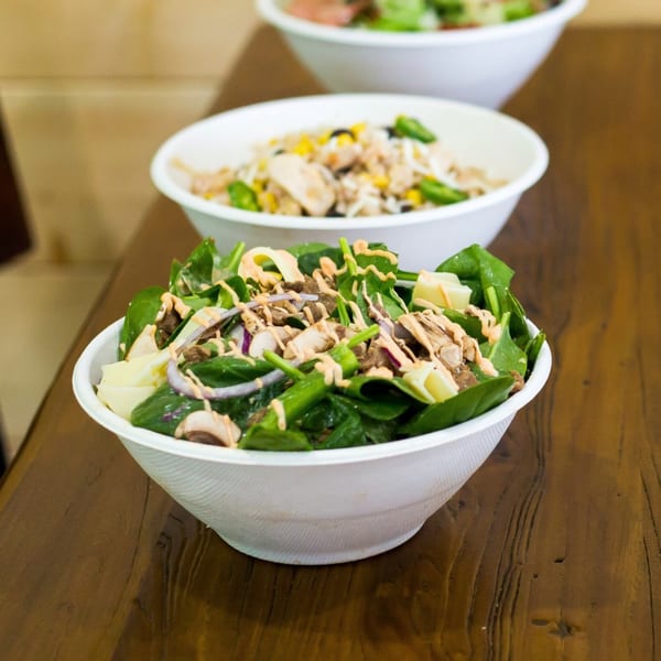 Three healthy Fuel Counter salads in white bowls on a wooden table, with spinach and dressing in the foreground.