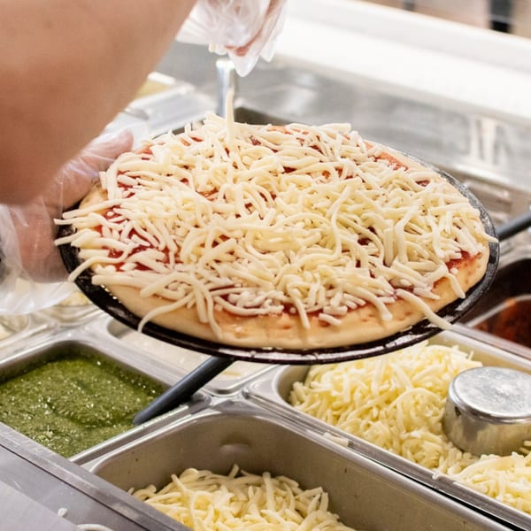 A gloved hand sprinkles shredded cheese onto a pizza with tomato sauce at Fuel Counter.