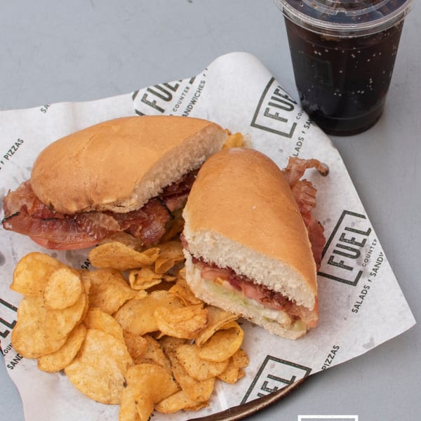 A Fuel Counter sandwich, chips, and a drink on branded paper, on a gray surface.