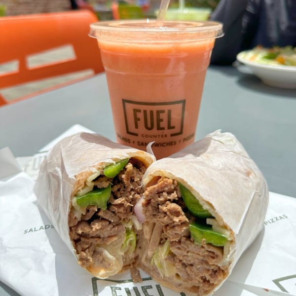 A Fuel Counter wrap, cut in half, filled with meat and green peppers, next to an orange smoothie.