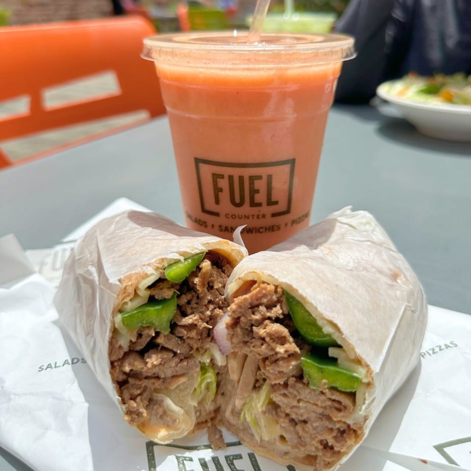 A Fuel Counter wrap, cut in half, filled with meat and green peppers, next to an orange smoothie.