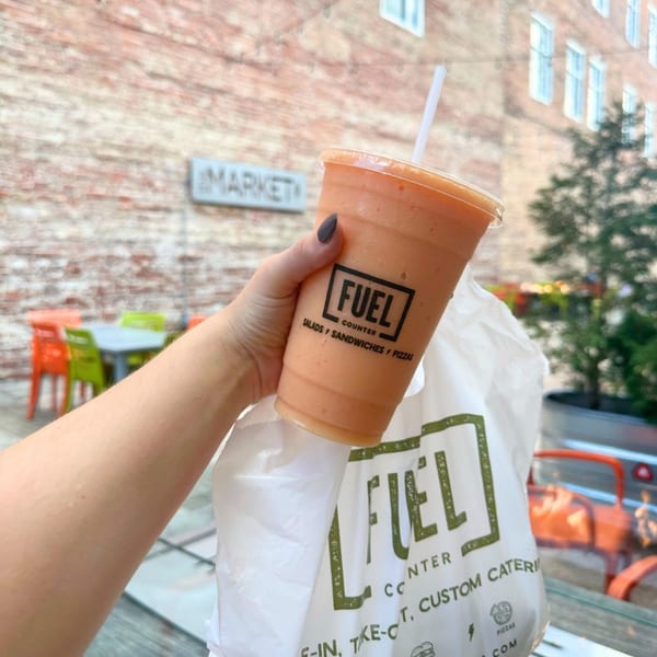 Hand holding a Fuel Counter smoothie and takeout bag outdoors, with brick buildings and market sign in background.